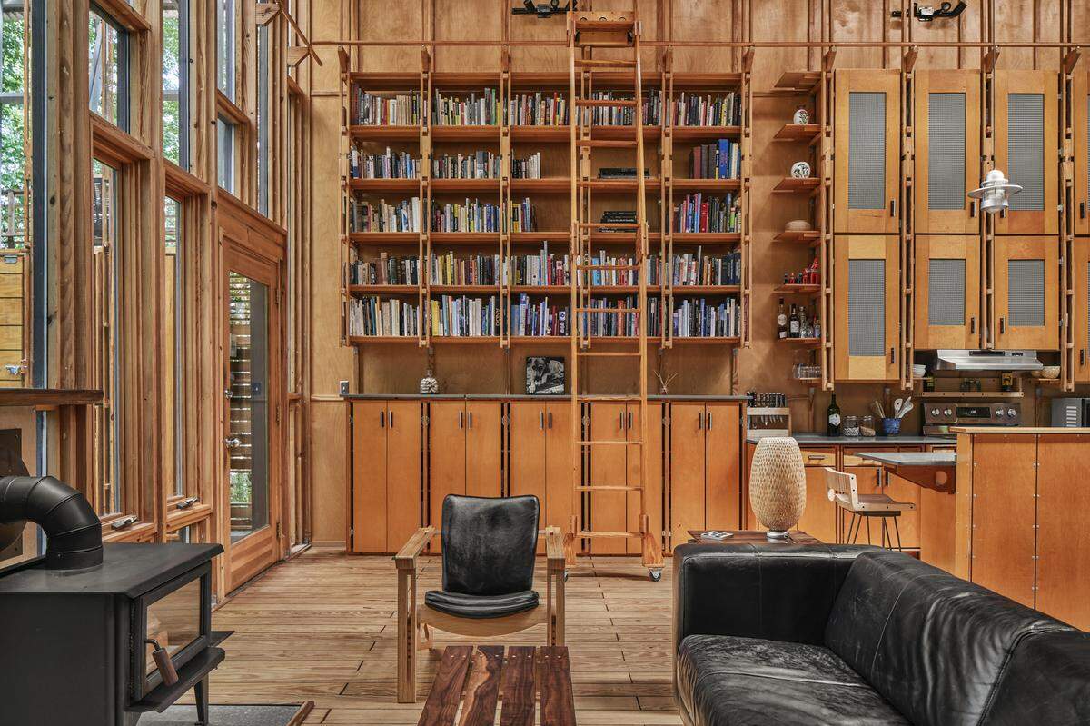 A wide, eye-level shot of a modern, wood-paneled cabin interior. The back wall features a floor-to-ceiling wooden bookshelf filled with books, complete with a rolling ladder. To the right is a kitchen area with matching wooden cabinets. In the foreground, a black leather sofa and a wood-framed armchair with black cushions make up a living space. On the far left, a black wood-burning stove is visible next to large, wood-framed glass windows and a door.