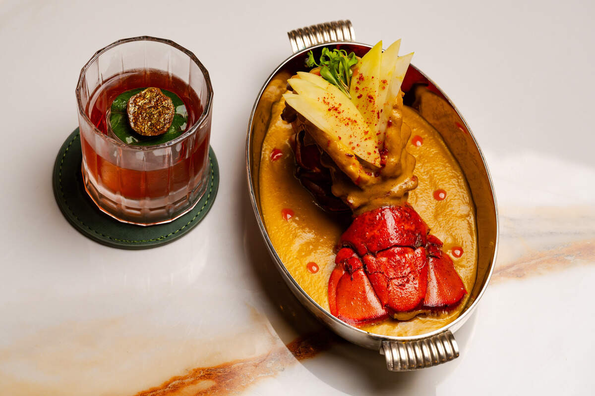 A cooked lobster tail is presented in an oval silver dish, coated in a rich golden curry sauce and garnished with thinly sliced apples and fresh microgreens. Next to the main dish, an amber-colored cocktail is served in a faceted lowball glass over a large ice cube topped with a green leaf and a piece of fig. The pair is neatly arranged on a clean, veined white marble countertop.