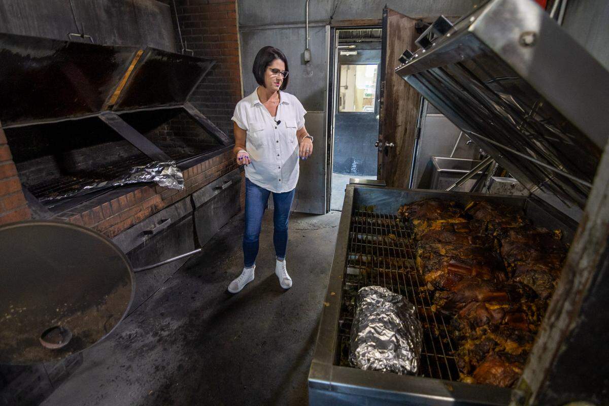 An indoor shot of a barbecue pit room, where a pitmaster with short dark hair and glasses, wearing a white shirt and jeans, stands between two large smokers. To the left is a built-in brick pit. To the right, the pitmaster is looking down into an open, stainless steel smoker filled with large cuts of cooked barbecue meat on a metal rack.