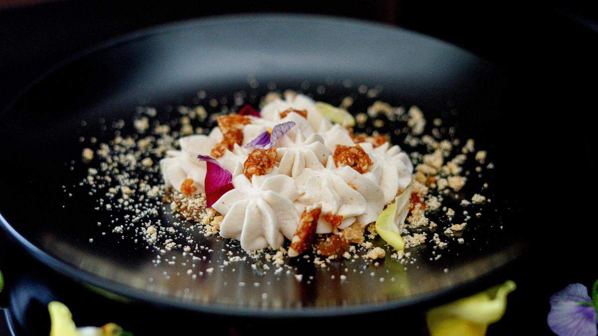 A close-up, low-angle shot of a dessert plated on a matte black dish. The dessert consists of a cluster of pale tan or off-white piped cream, possibly a mousse or whipped ricotta, surrounded by crumbles of toasted nuts or streusel, and small, reddish-brown pieces of candied ginger or fruit. It is garnished with two edible flowers: a dark magenta petal and a small purple and yellow pansy. The rest of the black plate and the background are dark, with a slight blur of yellow and green visible in the foreground.