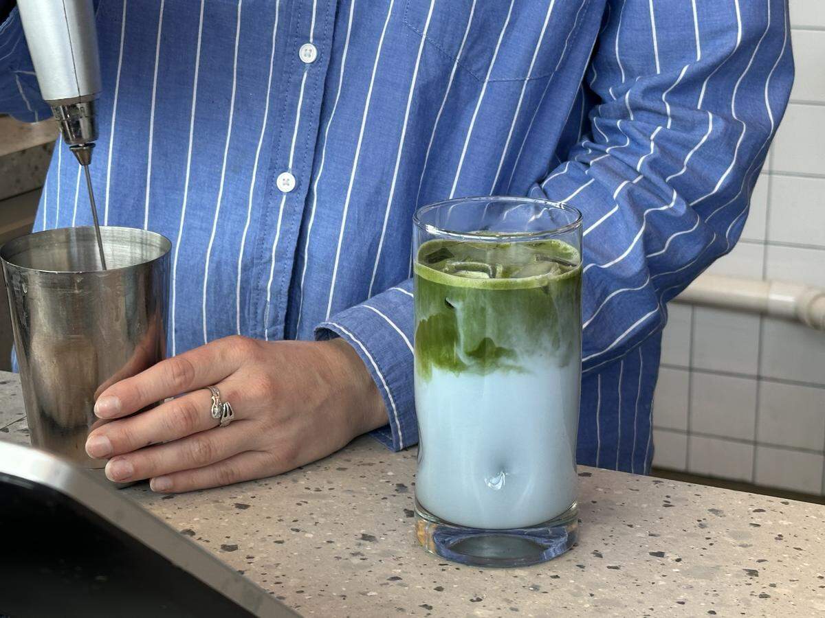 A close-up shot of a barista in a blue and white pinstriped shirt standing behind a speckled countertop. In front of them sits a tall glass with an iced, layered drink, featuring a light blue liquid at the bottom with a swirl of dark green matcha on top. To the left, the barista’s hand rests near a metal mixing cup, with an immersion blender held just above it.