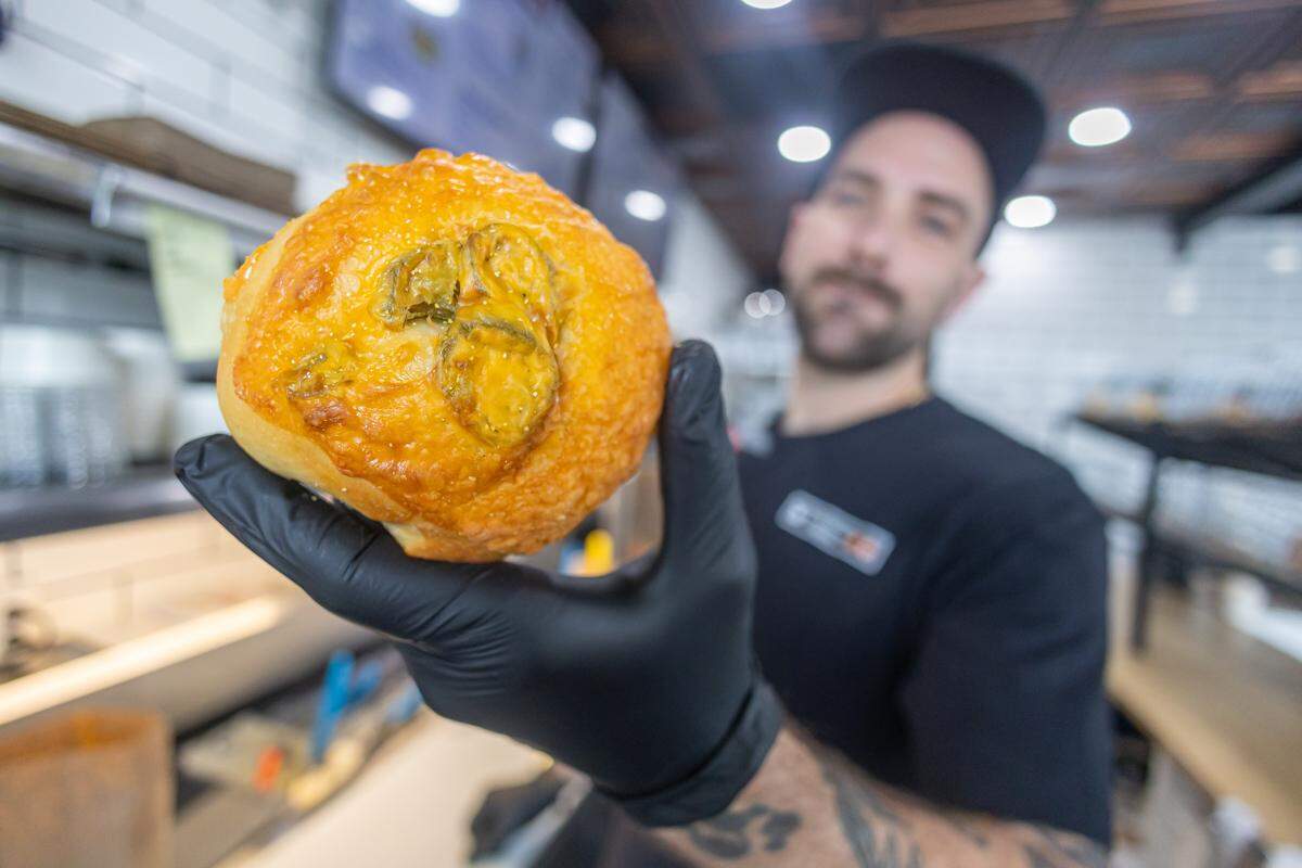 A baker, wearing a black glove, holds a freshly baked jalapeño cheddar roll close to the camera. The baker is slightly out of focus in the background of what appears to be a bakery.