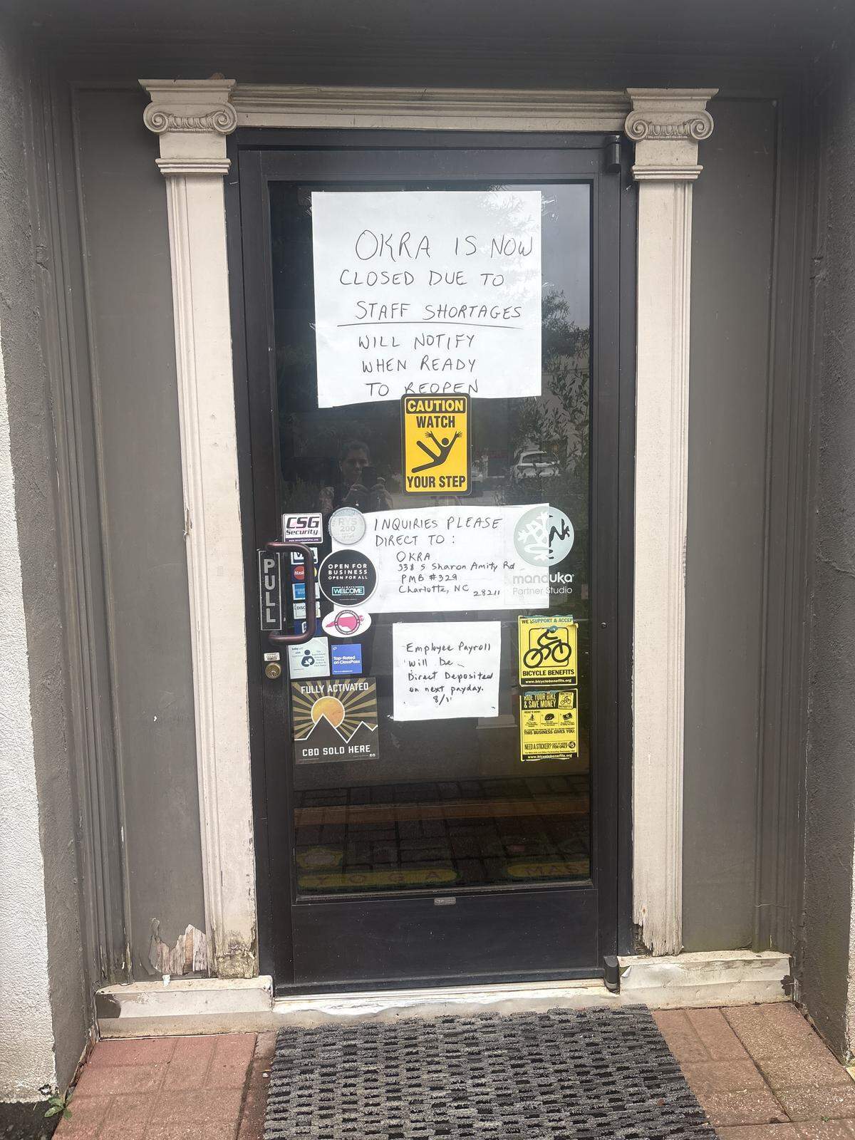 A straight-on, eye-level shot shows a glass door with a large white sign taped to it that reads, “OKRA IS NOW CLOSED DUE TO STAFF SHORTAGES. WILL NOTIFY WHEN READY TO REOPEN.” Below this, a smaller yellow sign with black text says “CAUTION WATCH YOUR STEP.” The door is framed by two decorative cream-colored columns, one on each side, with ornate capitals. Below the “OKRA” sign, several other smaller stickers and signs are affixed to the door, including “PULL” on a vertical black sticker, various logos, and a sign regarding “Employee Payroll.” A dark doormat with a textured surface lies on the ground in front of the door. The reflection of trees and a somewhat cloudy sky are visible in the glass. The building’s exterior walls on either side of the door are painted a neutral grayish-brown color.
