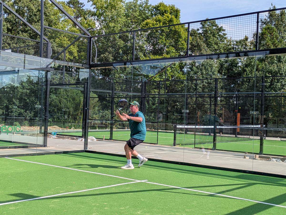 An action shot of a person playing padel on a sunny day, wearing a green t-shirt and black shorts. The player is positioned near the glass wall of the court, swinging a racket to connect with the ball on the backhand side.