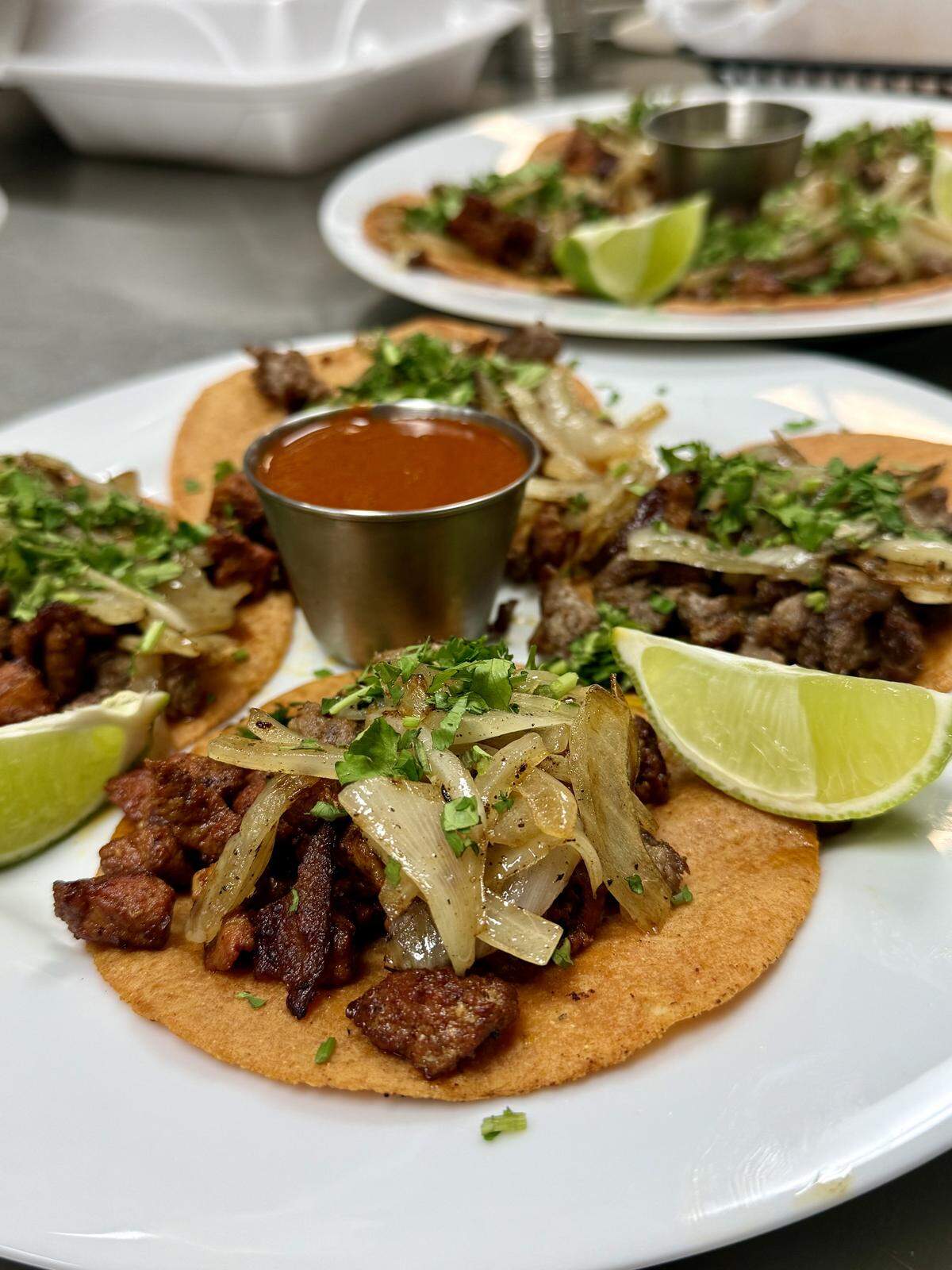 A white plate holding several street tacos filled with seasoned meat, grilled onions, and cilantro, with a cup of red salsa in the center and lime wedges on the side. Another plate of tacos is visible in the background.