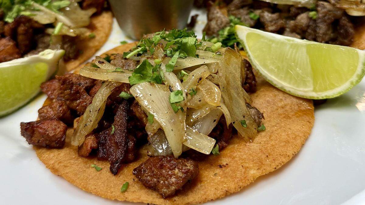A white plate holding several street tacos filled with seasoned meat, grilled onions, and cilantro, with a cup of red salsa in the center and lime wedges on the side. Another plate of tacos is visible in the background.