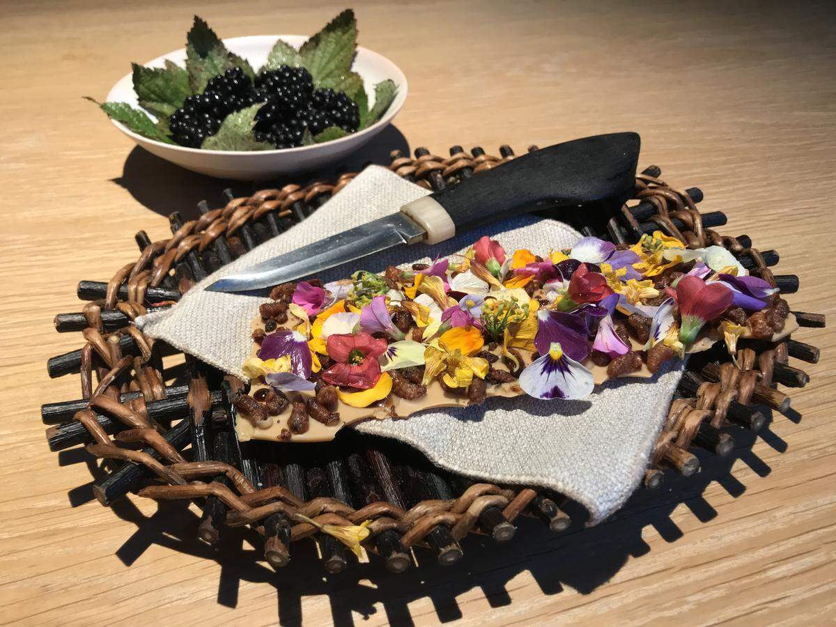 A slab of white chocolate topped with a dense layer of colorful edible flowers and candied nuts, presented on a linen napkin inside a rustic woven twig basket with a small knife.