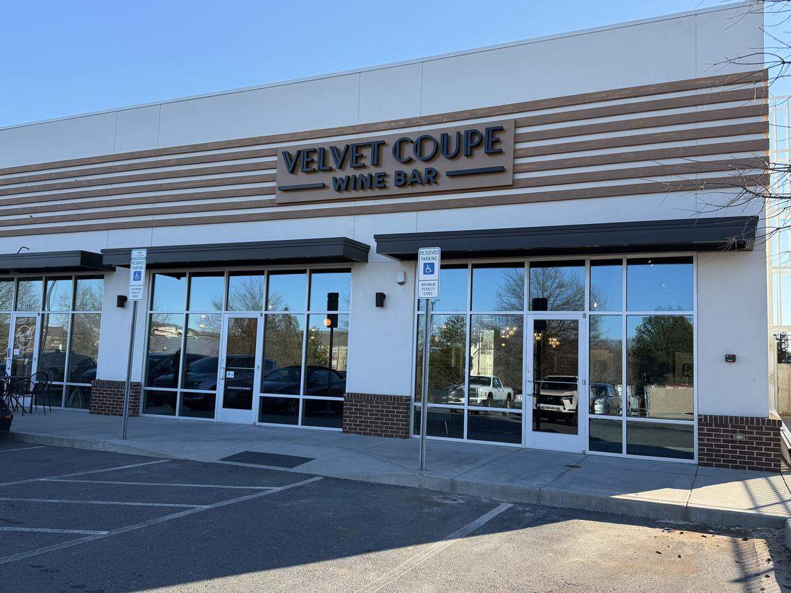 A daytime wide shot of the exterior storefront for “Velvet Coupe Wine Bar.” The building features a clean, white facade with horizontal wood-slat accents above the signage. Large floor-to-ceiling glass windows reflect a clear blue sky and a parking lot. Two black metal awnings hang over the entrances, and there are designated accessible parking signs in front of the brick-accented base of the building.