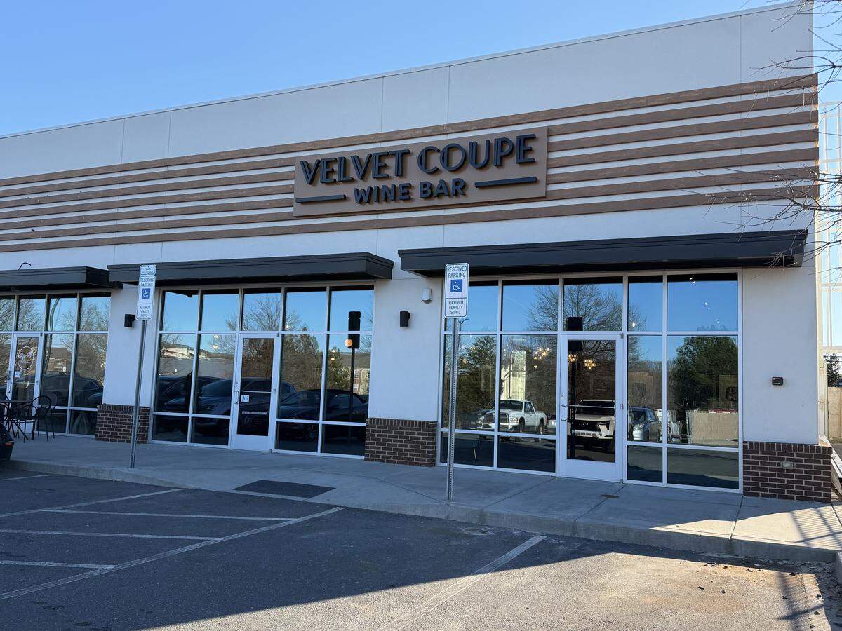 A daytime wide shot of the exterior storefront for “Velvet Coupe Wine Bar.” The building features a clean, white facade with horizontal wood-slat accents above the signage. Large floor-to-ceiling glass windows reflect a clear blue sky and a parking lot. Two black metal awnings hang over the entrances, and there are designated accessible parking signs in front of the brick-accented base of the building.