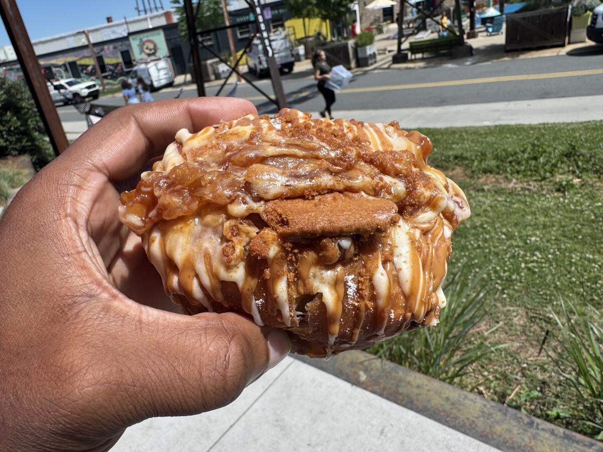 A person’s hand holds a large, decadent dessert eggroll outdoors. The eggroll is heavily drizzled with white icing and caramel sauce, topped with crushed cookie crumbles and a whole Biscoff cookie embedded in the center. The background shows a sunny street and green grass.