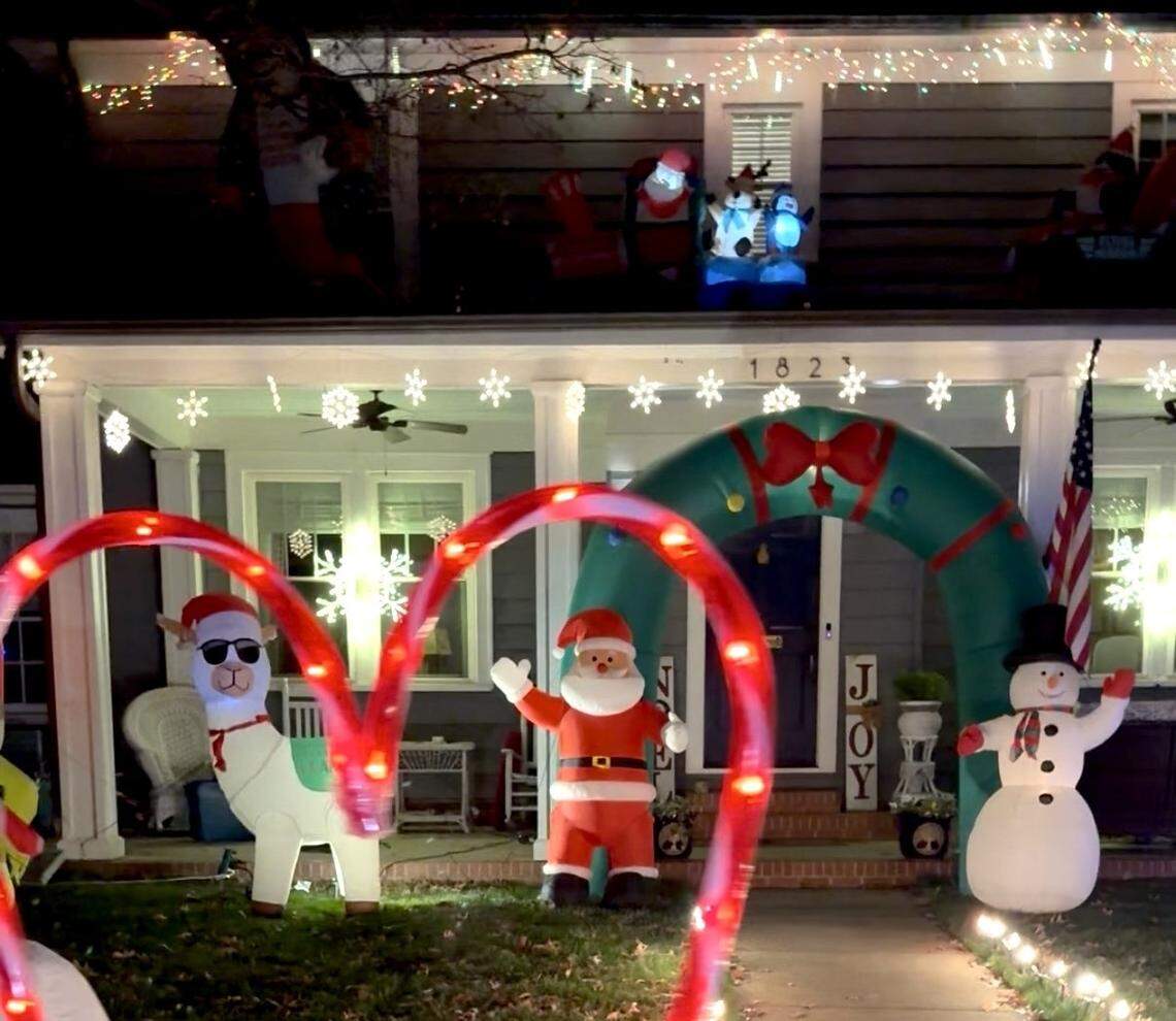 A close-up night view of a porch decorated with glowing snowflake lights and an address number ‘1823.’ The front lawn features a brightly lit scene of inflatables, including a large green wreath archway, a traditional Santa Claus, a cheerful snowman, and a comical llama figure. Red, curved candy cane lights border the front of the display.