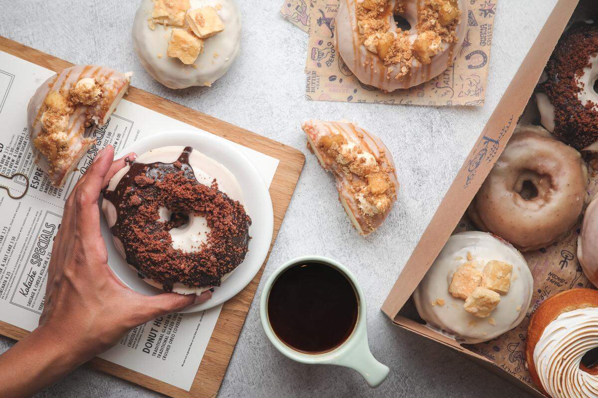 A top-down, overhead shot of an assortment of gourmet donuts and a cup of black coffee on a light grey textured surface. A hand reaches in from the left to pick up a donut topped with chocolate glaze and dark cookie crumbles. Several other donuts are visible, including some with white icing and square toppings, and others with caramel drizzle and crushed nuts.