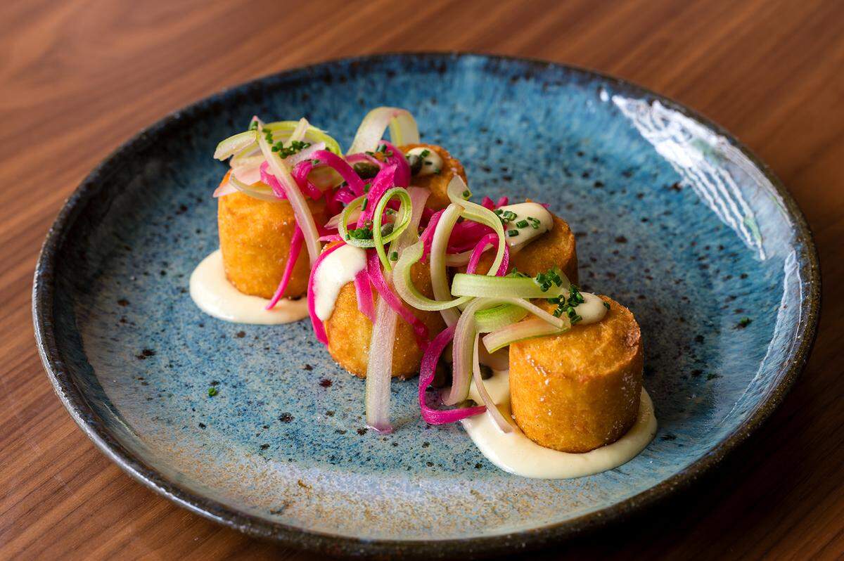 Three golden-brown, cylindrical potato croquettes or “tots” are artfully arranged on a blue ceramic plate with a textured, speckled finish. Each cylinder is topped with a dollop of creamy white sauce and a colorful tangle of pickled red onions, shaved green onions, and fresh chives. The plate sits on a polished wood tabletop, emphasizing a modern, gourmet culinary style.