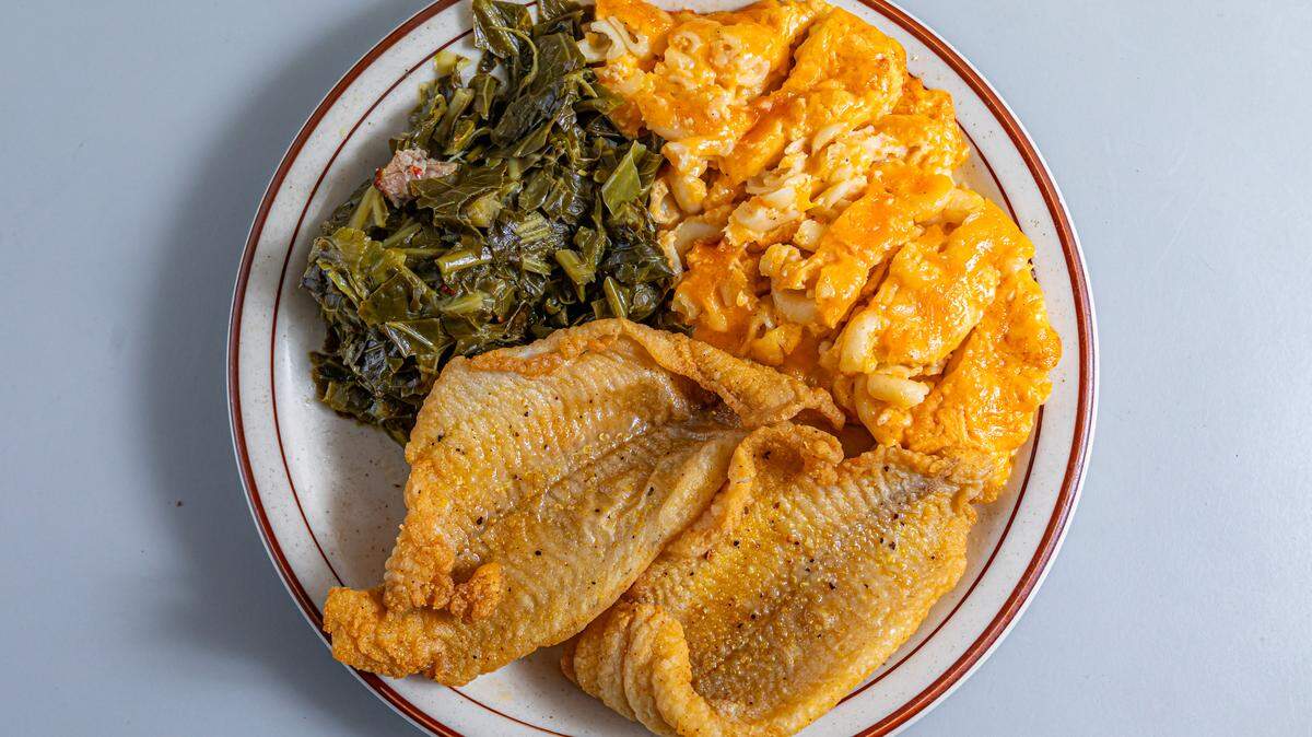 A top-down view of a plate filled with a traditional soul food meal. In the foreground, two pieces of golden-brown fried fish fillets are lightly seasoned with black pepper. The back left of the white, brown-rimmed plate features a generous serving of dark green, slow-cooked collard greens, while the back right holds a large portion of baked macaroni and cheese with a melted orange cheese crust.