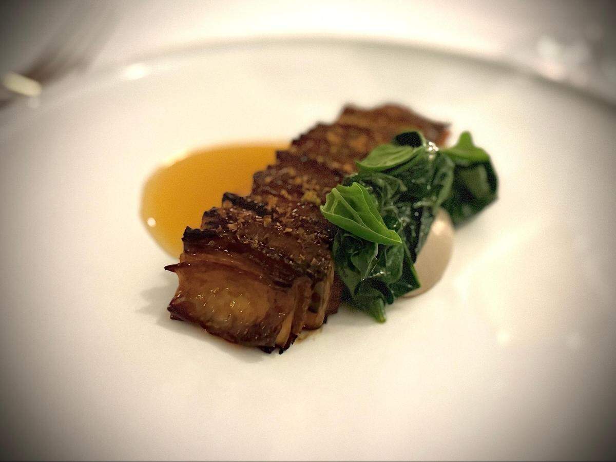 A close-up, angled shot of a meticulously plated dish on a wide, white rimmed plate. The main element is a rectangular portion of braised or roasted meat or mushroom, sliced into several pieces, fanned out, and resting in a small pool of amber-colored sauce. Beside it is a neat pile of bright green, wilted spinach.