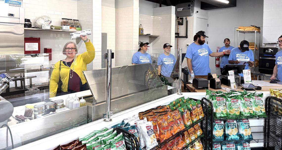 A bright, modern deli counter at Ricci’s. Behind a glass partition and metal prep station, several staff members in matching light blue t-shirts work near a digital register and a rack of fresh long rolls. In the foreground, wire racks are filled with various bags of Herr’s potato chips. A woman in a yellow sweater stands at the counter holding up a wrapped sandwich.