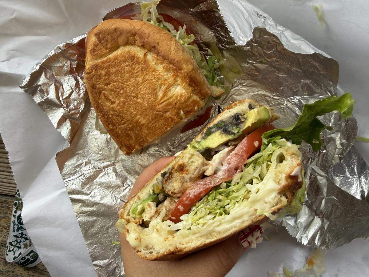 A close-up, overhead shot of a person holding half of a substantial sandwich on a crusty roll. The cross-section reveals layers of grilled chicken, sliced avocado, tomato, shredded lettuce, and a creamy sauce. The other half of the sandwich lies on the open foil and paper wrapper.
