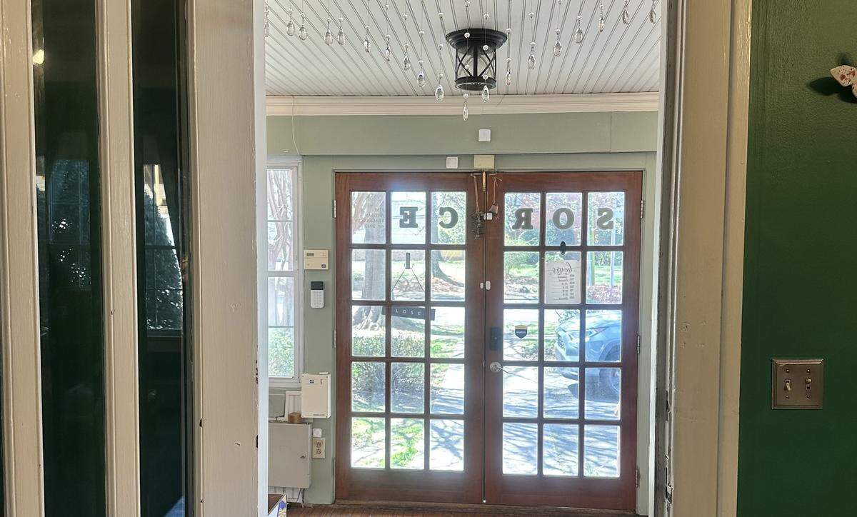 A view from inside a building looking out through a pair of wood-framed French doors. Above the doors, a decorative beaded curtain hangs from the ceiling around a black, lantern-style light fixture. Through the glass, a residential street with trees and a blue car is visible. The interior walls are a soft sage green.