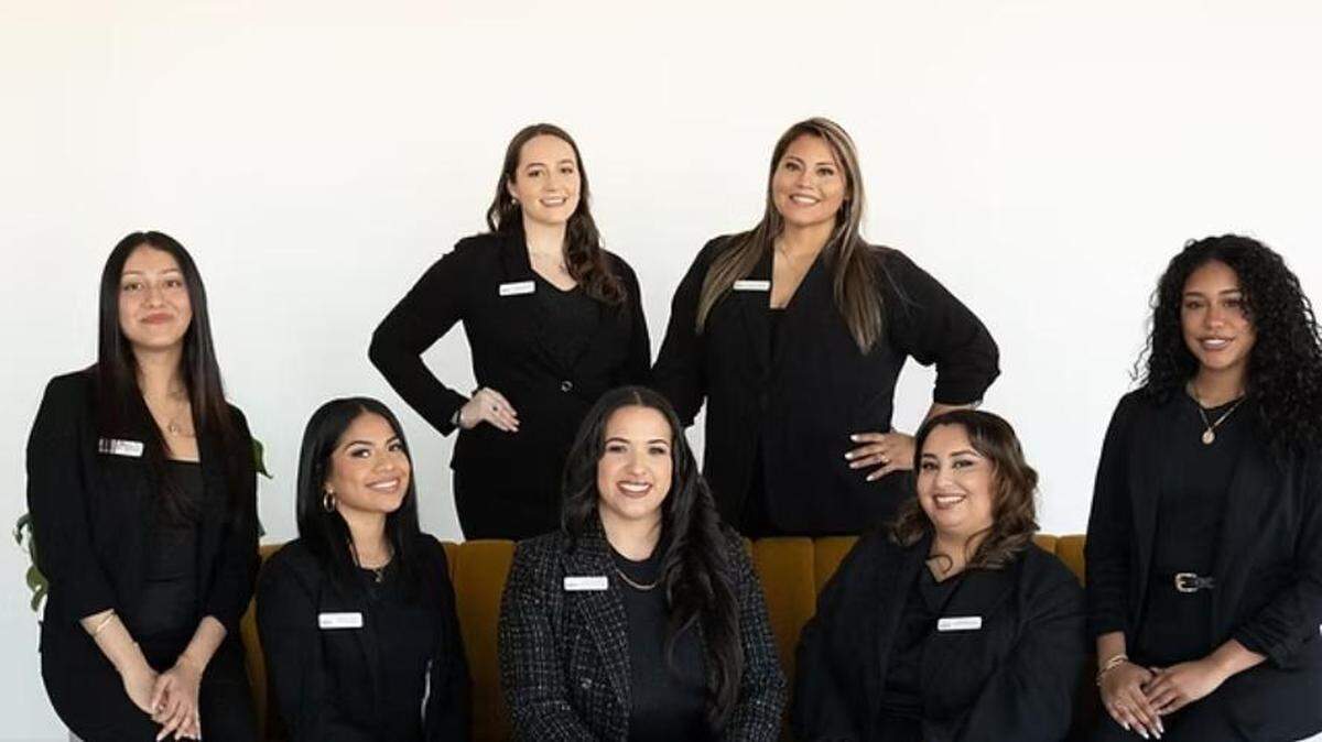 A group studio portrait of seven people, all smiling and wearing black professional attire. Four stand in the back, while three are seated in the front on an upholstered yellow bench against a solid white background.
