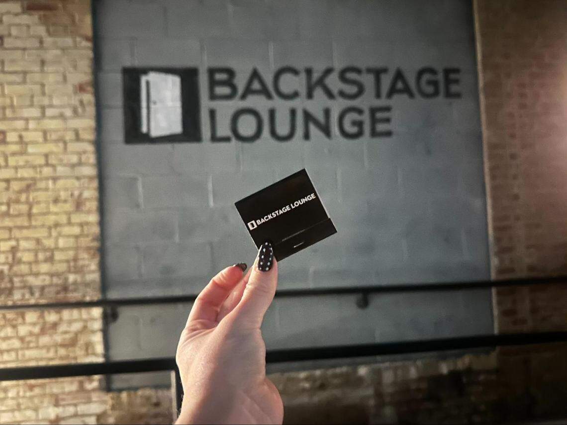 A person’s hand, with a black and white polka-dotted manicure, holds up a small, black “BACKSTAGE LOUNGE” pass. The pass is held in front of a matching, larger sign painted on a gray cinder block wall, creating a layered effect. An exposed brick wall is visible on the left side of the frame.