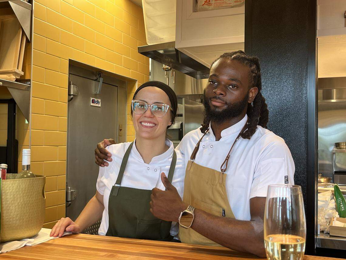 A portrait of a smiling man and woman standing behind a wooden counter in a professional kitchen. The woman on the left, wearing glasses, a black head covering, and a green apron over her white chef’s jacket, is giving a thumbs-up. The man on the right, with dark hair in dreadlocks, is wearing a white chef’s jacket with a brown apron and has his arm around the woman’s shoulder. They are both looking directly at the camera. A glass of white wine is visible on the counter in the foreground. The kitchen has bright yellow tile walls and stainless steel appliances.