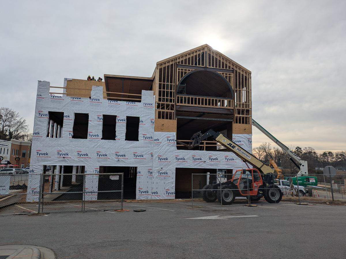 A large multi-story building under construction with wooden framing and a gabled roofline. Much of the exterior is covered in white Tyvek commercial wrap, and several workers are visible on an upper level. In the foreground, an orange JLG telehandler and other construction equipment are parked behind a chain-link fence on an asphalt lot under an overcast sky.