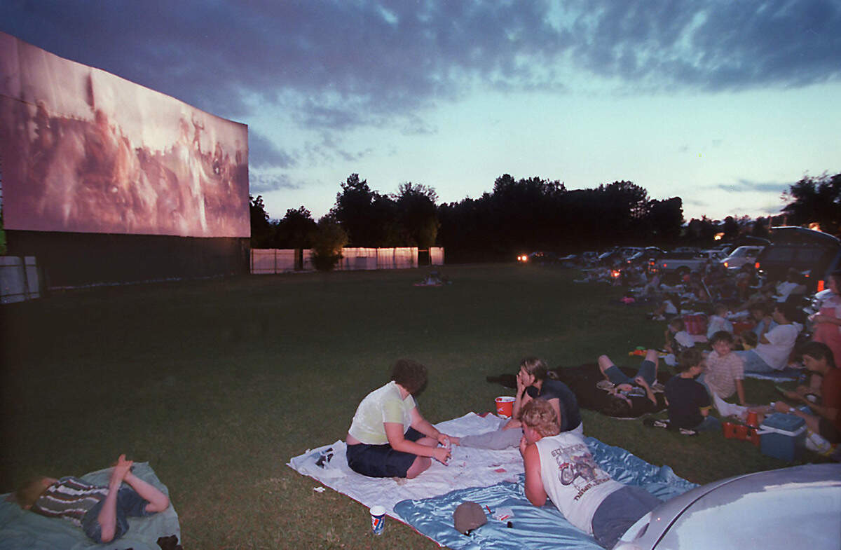A wide shot of a drive-in movie theater at dusk. On the far left, a large outdoor screen is illuminated, showing a movie to a crowd spread across a large grassy field. Many people are watching from blankets, lawn chairs, and parked cars. In the foreground, a small group sits on a blue and white blanket on the grass, while another child lies on a blanket to their left. The background is lined with dark trees under a cloudy, twilight sky.
