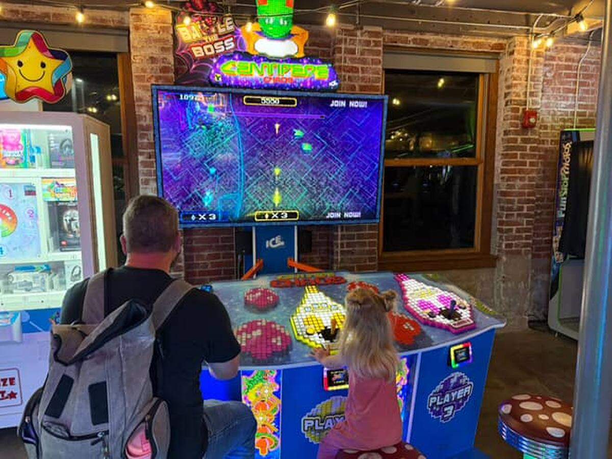 A father and young daughter are fully engaged in playing a Centipede-themed arcade game together. The girl sits on a spotted mushroom stool, while the father has a backpack on, with the bright blue machine and large screen dominating the center of the frame.