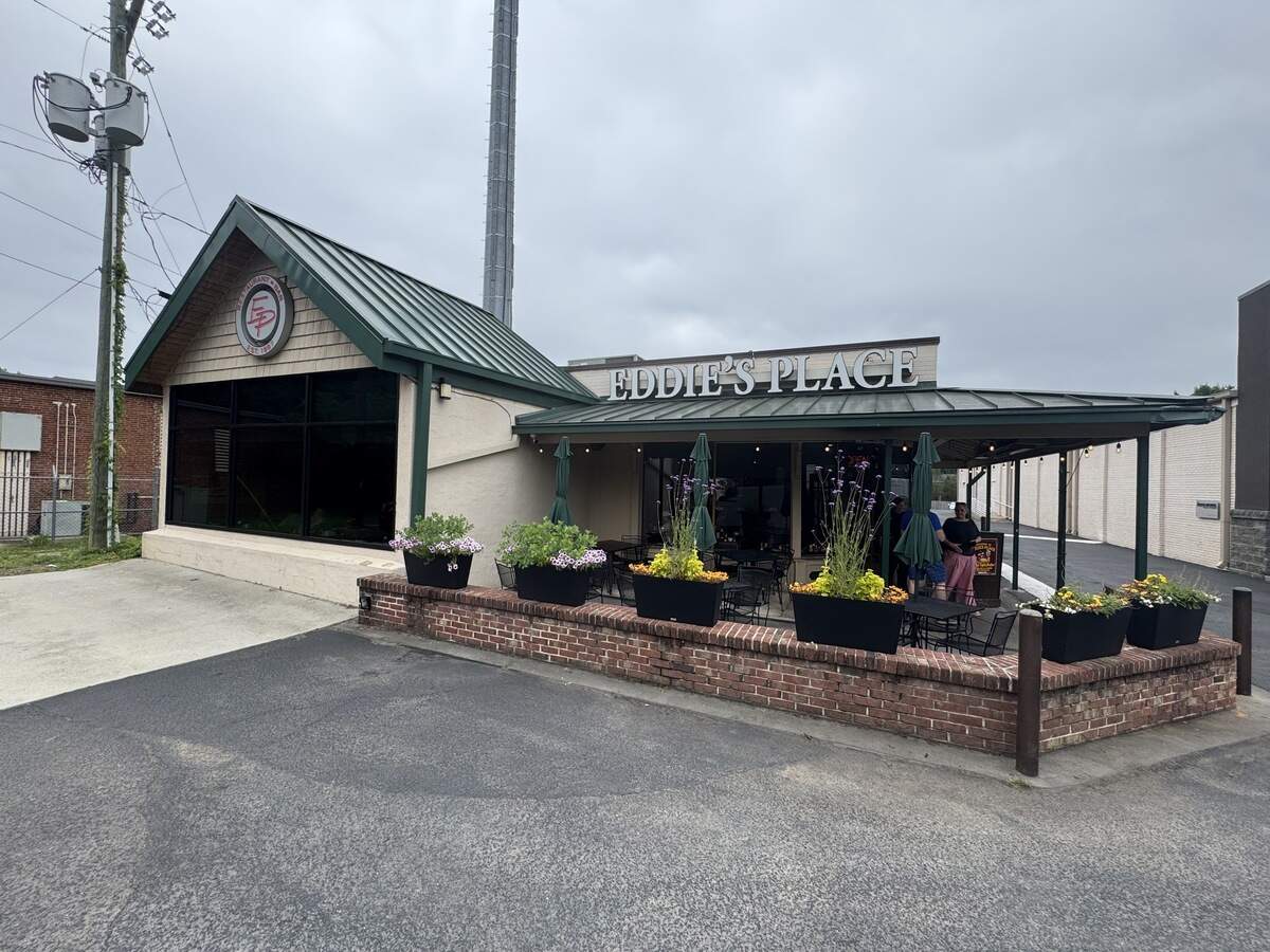 The exterior storefront of Eddie’s Place Restaurant and Bar under an overcast sky. The building features a green metal roof, cream-colored walls, and a small covered patio area with outdoor seating. A low brick wall lined with black rectangular planters full of flowers separates the patio from the asphalt parking lot in the foreground.