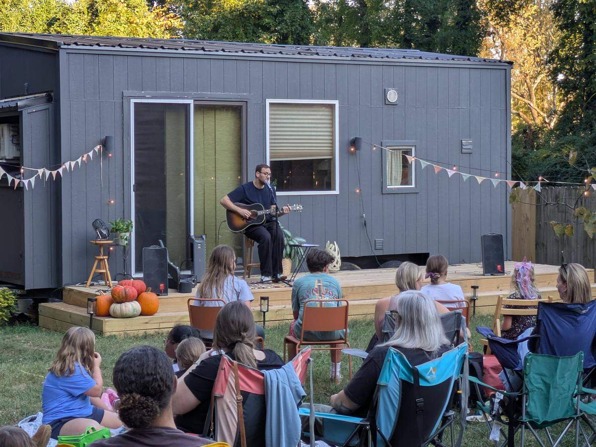 A wide shot of an intimate backyard concert, where a man sits on a stool on a low wooden deck, playing an acoustic guitar. He is performing in front of a gray tiny house, with speakers, a string of pennant flags, and pumpkins decorating the small “stage.” An audience of adults and children, seen from behind, sits in camping chairs on the grass, watching the performance.