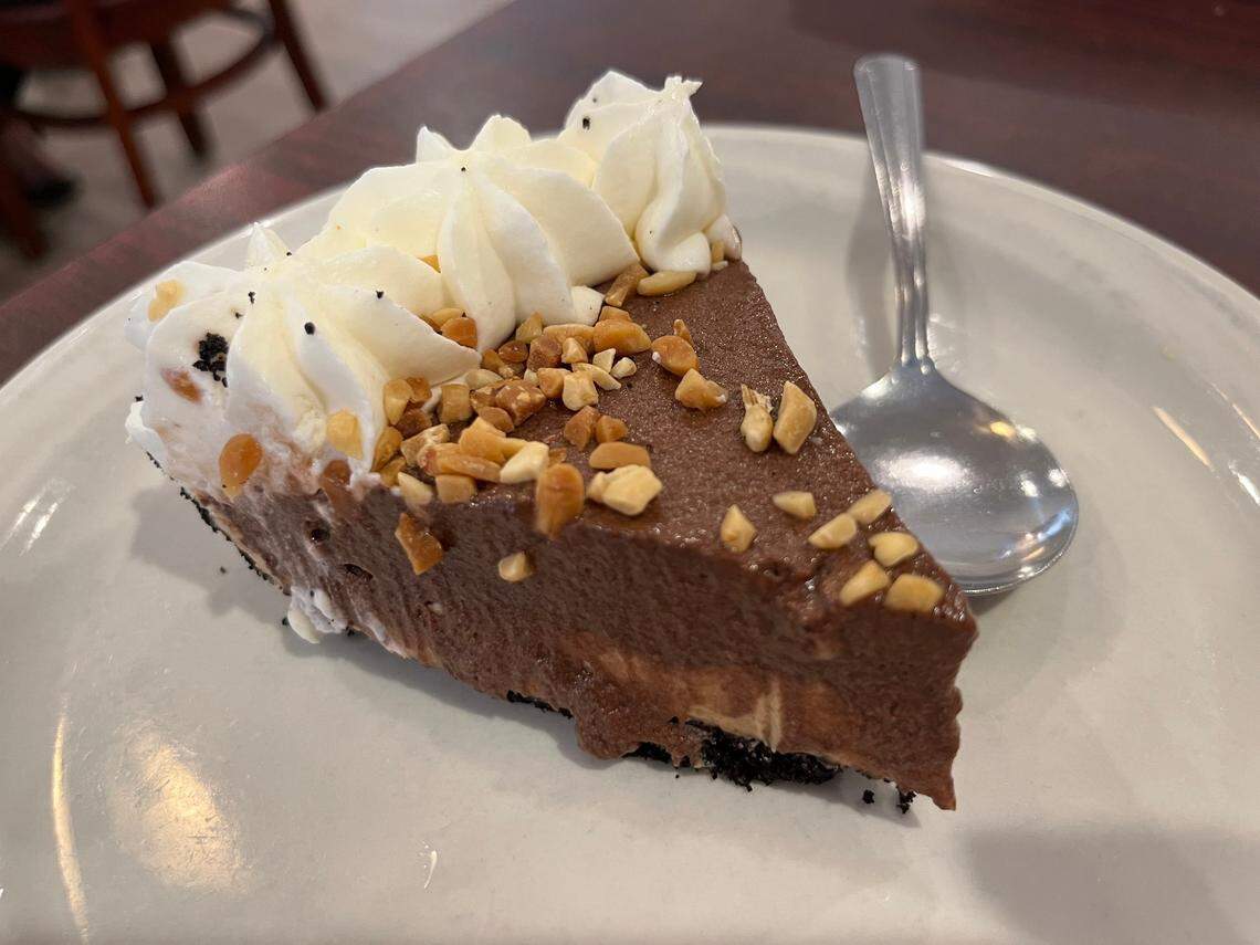 A wedge of rich chocolate cream pie with a dark crust sits on a white plate, topped with generous swirls of whipped cream and a sprinkle of chopped peanuts. A silver spoon rests next to the slice, ready to scoop the dessert.
