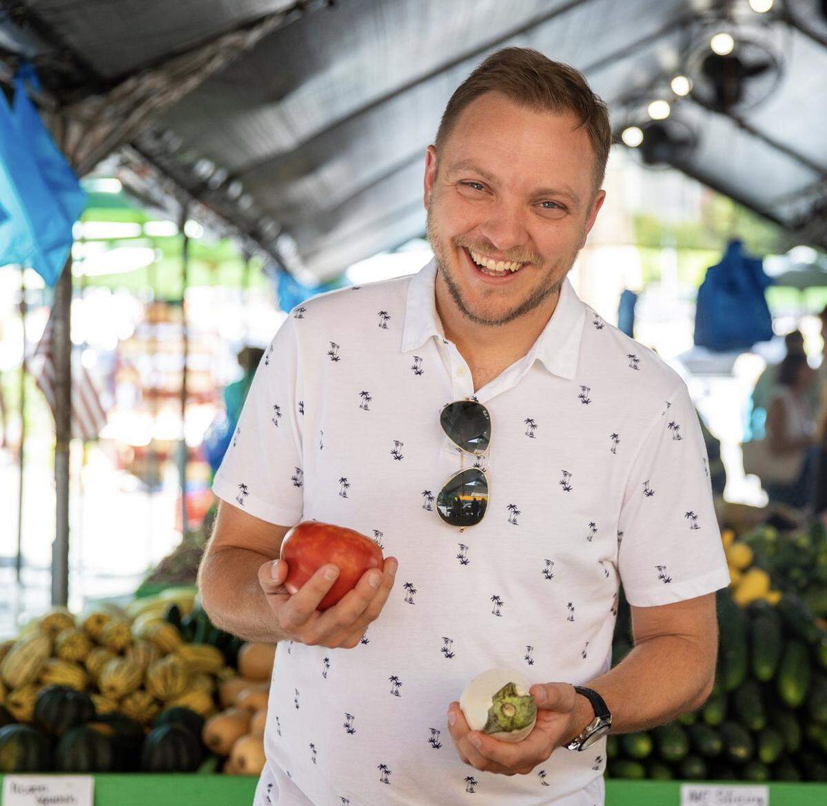 A smiling man with light hair and a goatee stands in a bustling outdoor market, wearing a white polo shirt with a palm tree pattern and sunglasses tucked into his collar. He holds a large, ripe red tomato in his right hand and a small, round white eggplant in his left. Behind him, crates are overflowing with fresh produce, including squash and cucumbers, under a shaded canopy.