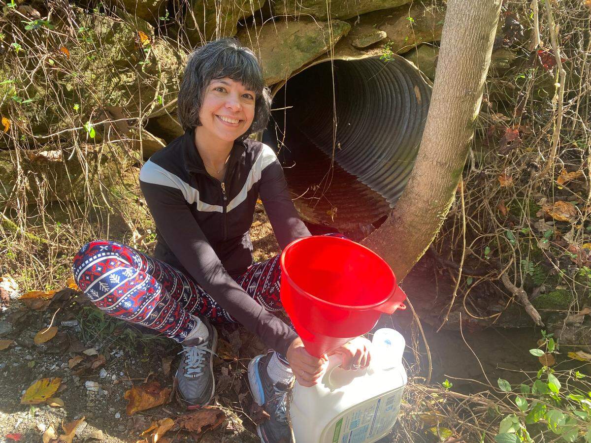 On a sunny day in a rustic, wooded setting, a person crouches next to a creek, smiling and looking at the camera while holding a large red funnel to guide water from a culvert pipe into a gallon jug.