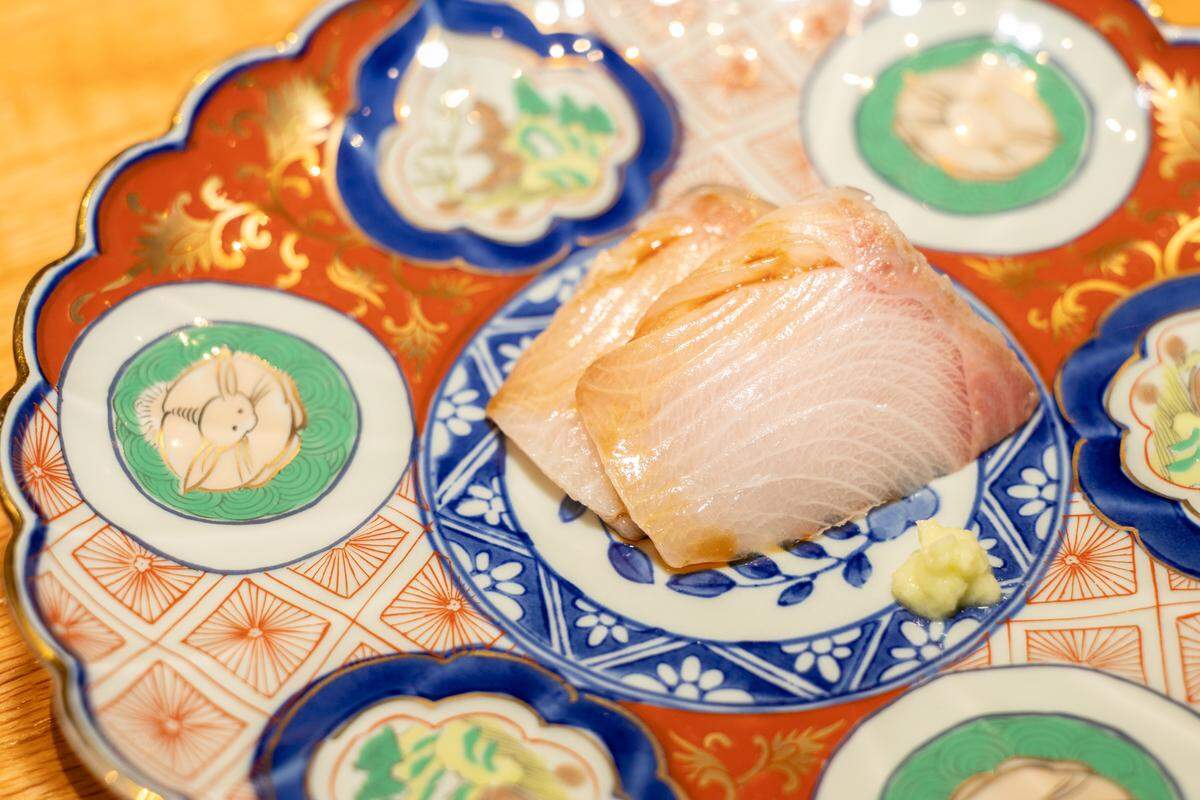 A close-up, high-angle shot of two slices of pale, translucent white fish sashimi served on a decorative, multi-colored ceramic plate with blue, red, and green patterns. A small dollop of pale green wasabi sits to the side.