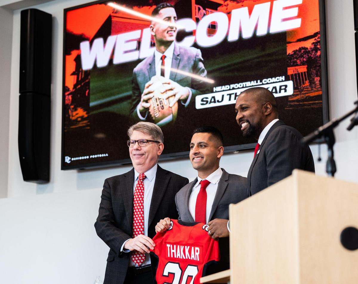 Davidson College president Douglas Hicks, left, and Director of Athletics Chris Clunie stand next to Saj Thakkar, center, as he holds up a jersey after being introduced as the new football coach for Davidson College in Davidson, N.C., on Friday, December 20, 2024.