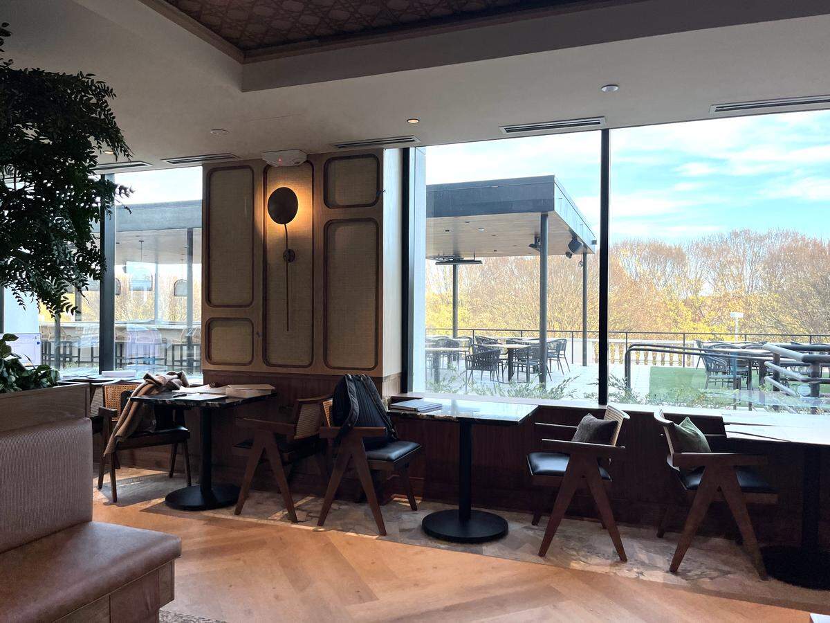 A wide, eye-level shot of a modern restaurant dining room featuring small tables and dark wooden chairs positioned by large, floor-to-ceiling windows. The view outside shows a spacious patio with covered seating and a backdrop of trees with autumn foliage under a bright sky.
