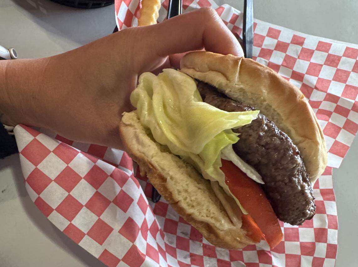A handheld view of a burger from the Park Road Soda Shoppe in Charlotte, a classic diner known for its hand-patted 80/20 chuck burgers. The burger is wrapped in red-and-white checkered paper and topped with fresh leaf lettuce and a tomato slice.