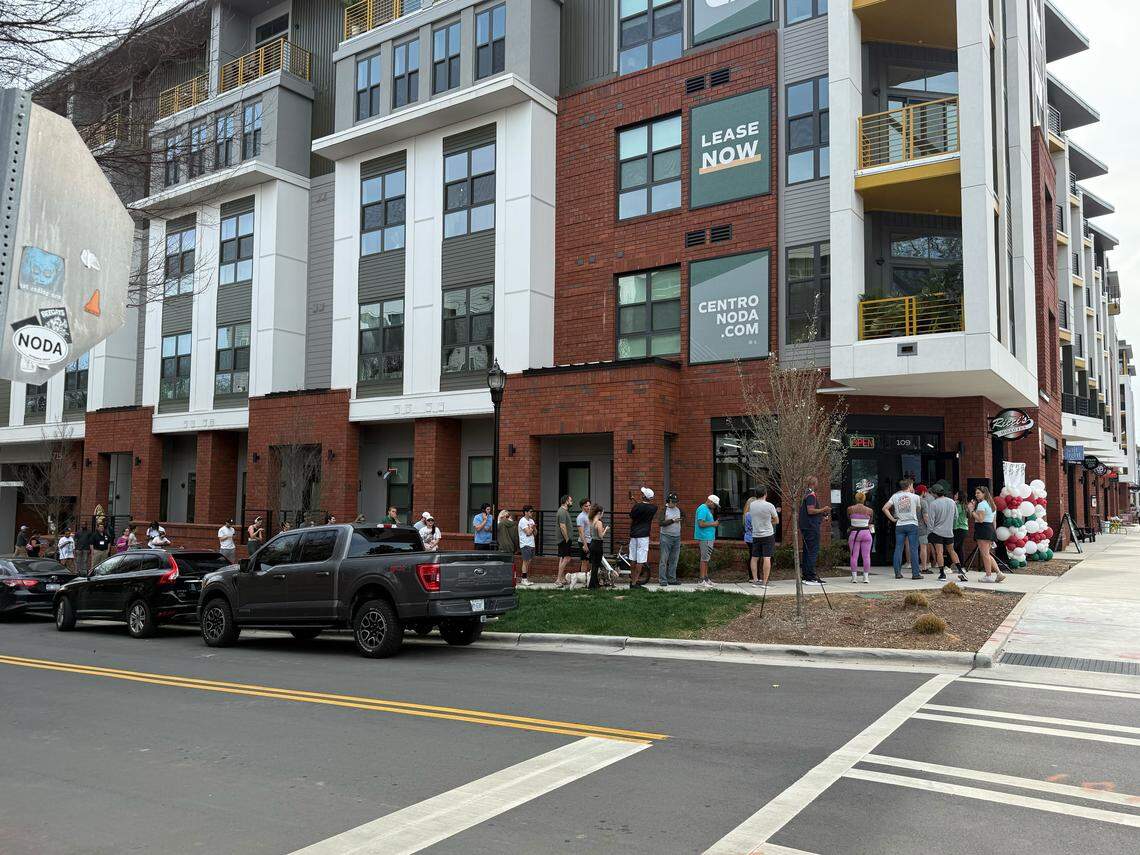 A street-level shot of a long queue of people. The focus is on the crowd waiting along the brick exterior of the building. A street sign in the far left foreground features various stickers, including one that says “NoDa.”
