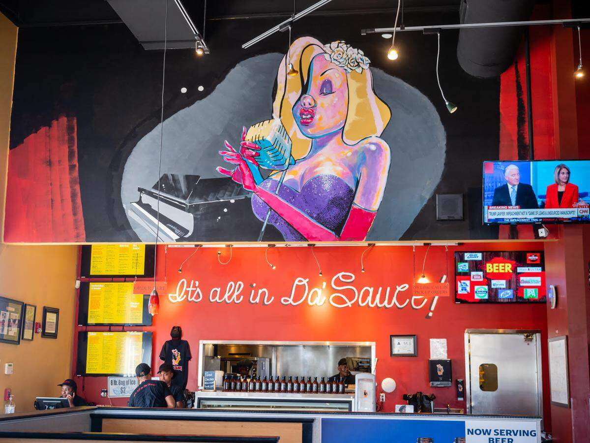 A wide shot of a restaurant interior featuring a large, colorful mural of a woman singing into a vintage microphone. Below the mural, a bright red wall is decorated with white cursive text that reads “It’s all in Da’ Sauce!” and a line of bottled sauces on a shelf.