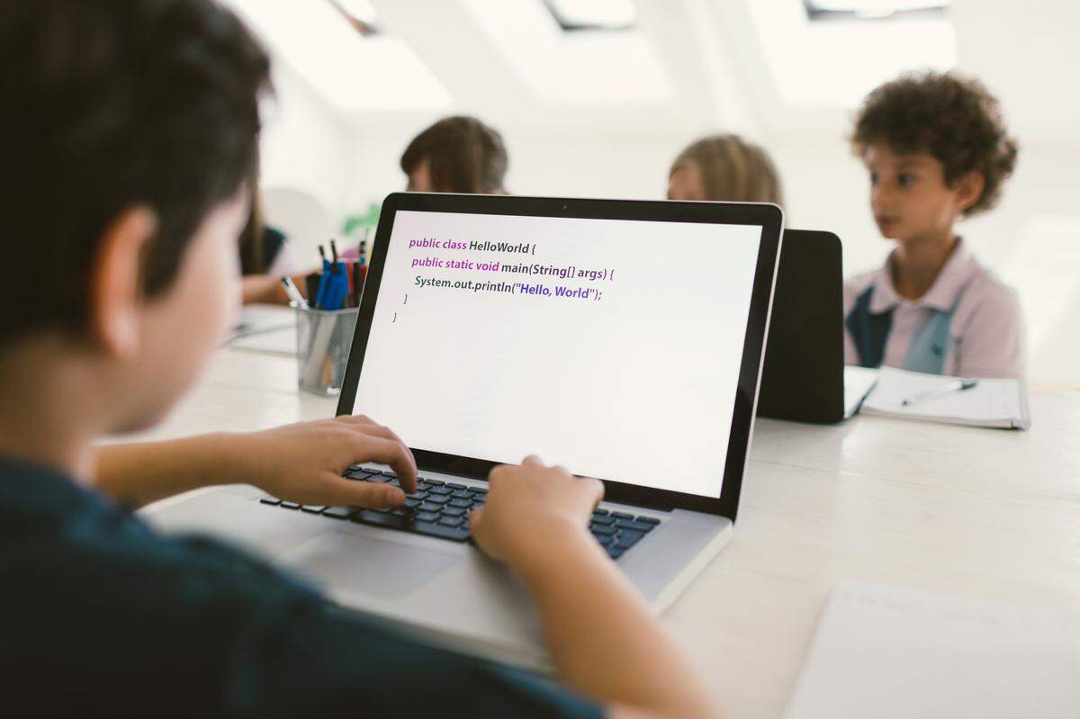 10 years old boy at coding class. He is sitting by the desk and using laptop, coding. He is focus on his work. Kids in background sitting and coding. Selective focus on laptop monitor.
