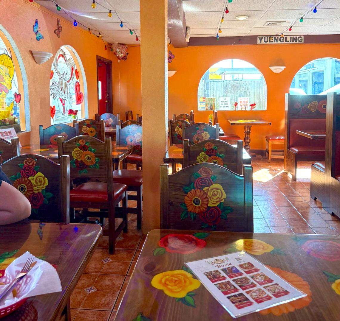 The interior of a restaurant features vibrant orange walls and multiple arched windows alongside multi-colored string lights draped across the ceiling. Hand-painted red and yellow floral designs decorate the wooden chair backs and tabletops, and a Birria-themed menu rests on the table in the foreground.