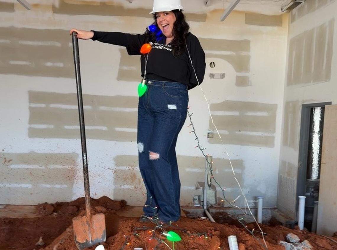 A full-length shot of a person with long dark hair, wearing a white hard hat, a black long-sleeved shirt, and ripped blue jeans, standing on a pile of dirt inside a room under construction, holding a shovel with one hand and smiling enthusiastically. The person is draped in a string of large, colorful Christmas lights that wrap around the torso and trail down onto the dirt. The background shows unfinished white walls with patches of brown plaster.
