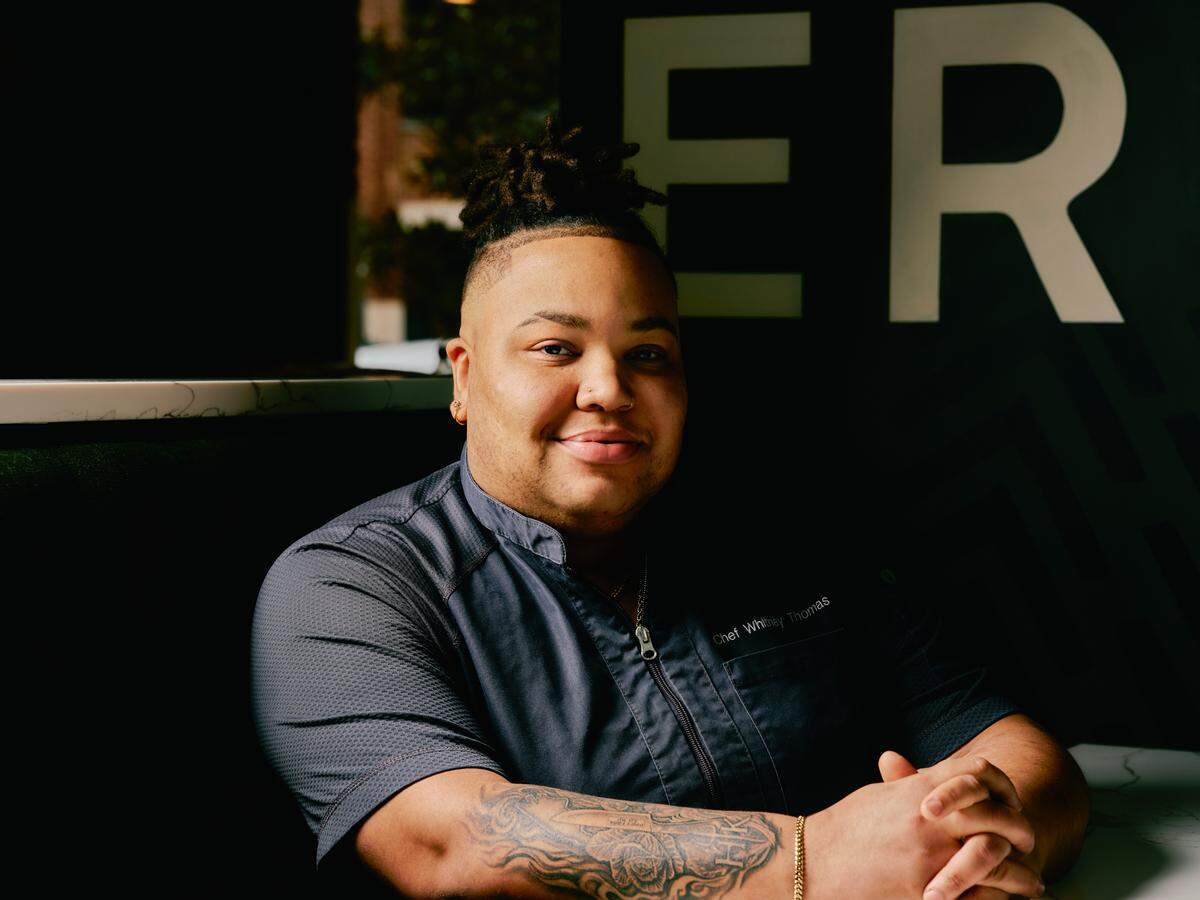 A warmly lit portrait of a chef smiling while sitting in a restaurant booth. The chef is wearing a dark gray chef’s coat, and tattoos are visible on her left forearm. In the dark background behind, the large white letters “ER” are visible on a wall.
