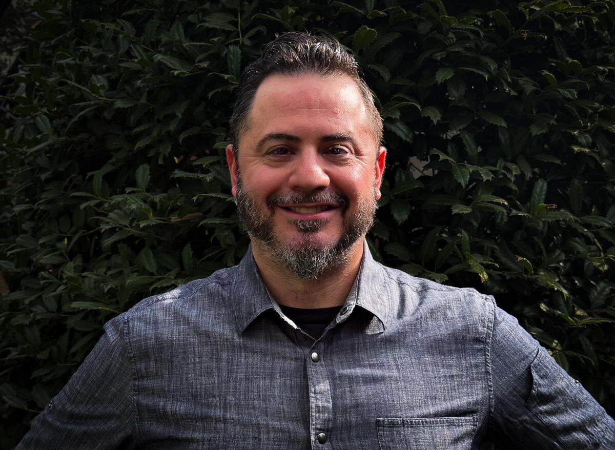 A professional, chest-up portrait of Chef Brett Faldstein. He is a smiling man with light eyes and a neatly groomed beard, looking directly at the camera. He is wearing a grey textured shirt, and the background is a solid, dark green wall of foliage.