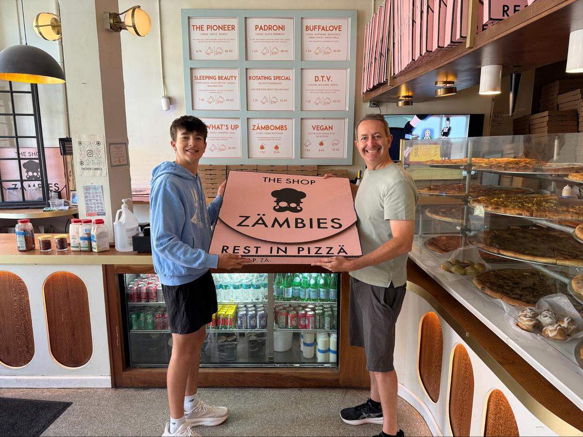 A wide, eye-level shot inside a pizzeria showing a smiling teenager and another person proudly holding a massive, oversized pink pizza box together. The teenager on the left wears a light blue hoodie, black athletic shorts, and white sneakers, while the person on the right wears a plain light gray t-shirt, dark gray shorts and black athletic shoes. The large pink box features a skull logo with a chef’s hat and reads “THE SHOP ZÄMBIES” and “REST IN PIZZA.”