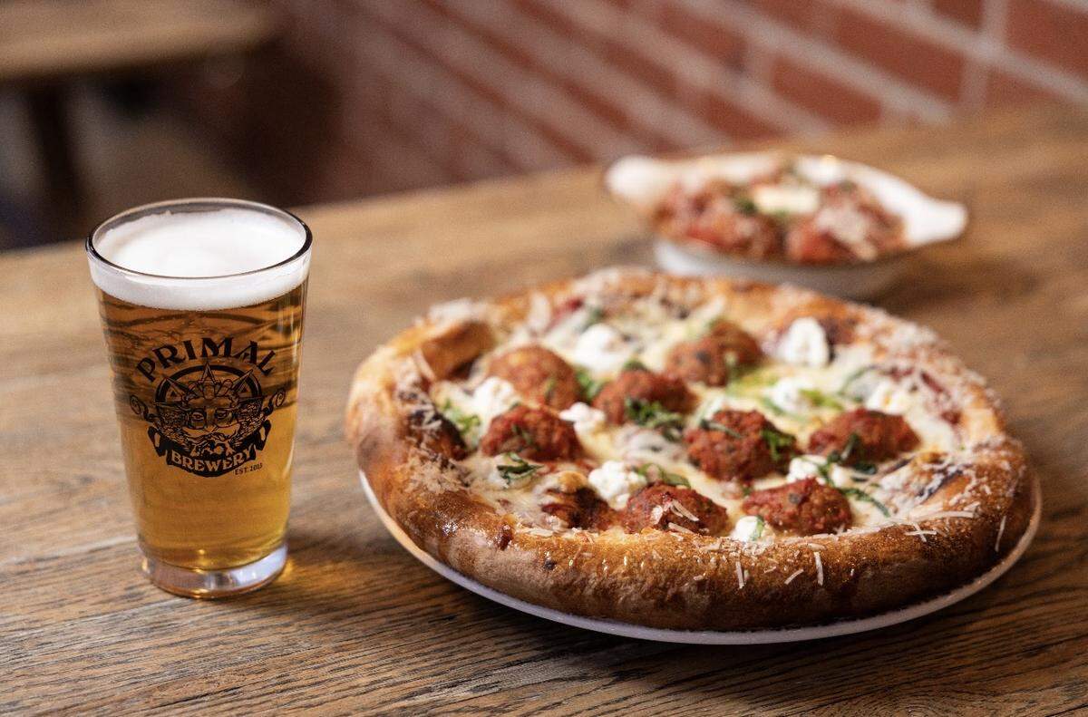 A golden pint of Primal Brewery beer stands next to a wood-fired meatball pizza topped with fresh basil and dollops of white cheese. In the background, a small white dish holds extra meatballs in marinara sauce, all set against a rustic brick wall.