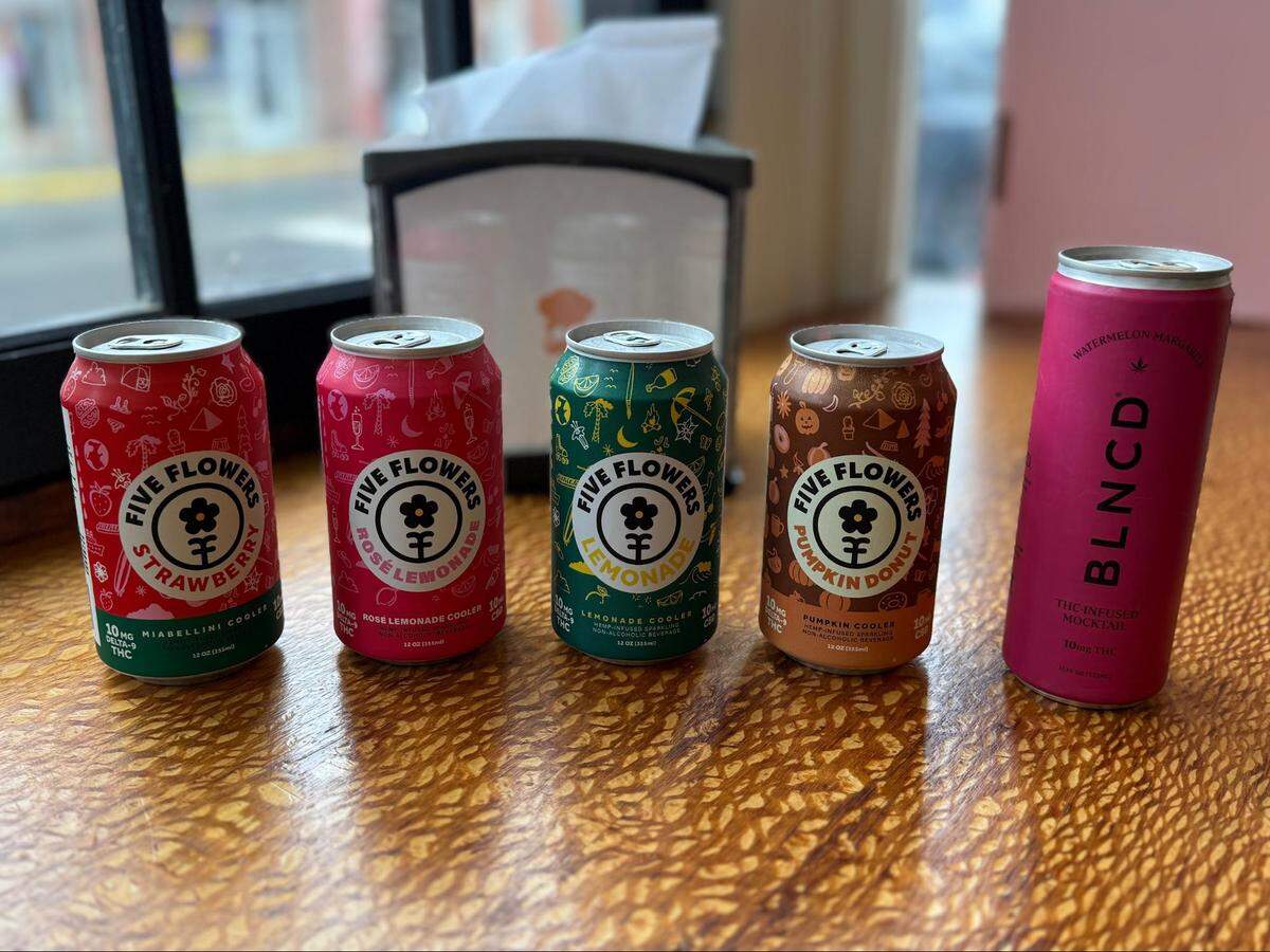 A close-up, eye-level shot of five colorful cans of THC-infused beverages lined up on a wooden table with a distinctive grain pattern. In the background, a napkin dispenser and a window are blurred. From left to right, the cans are: “Five Flowers Strawberry,” “Five Flowers Rosé Lemonade,” “Five Flowers Lemonade,” “Five Flowers Pumpkin Donut,” and a taller, pink “BLNCD Watermelon Margarita” mocktail.