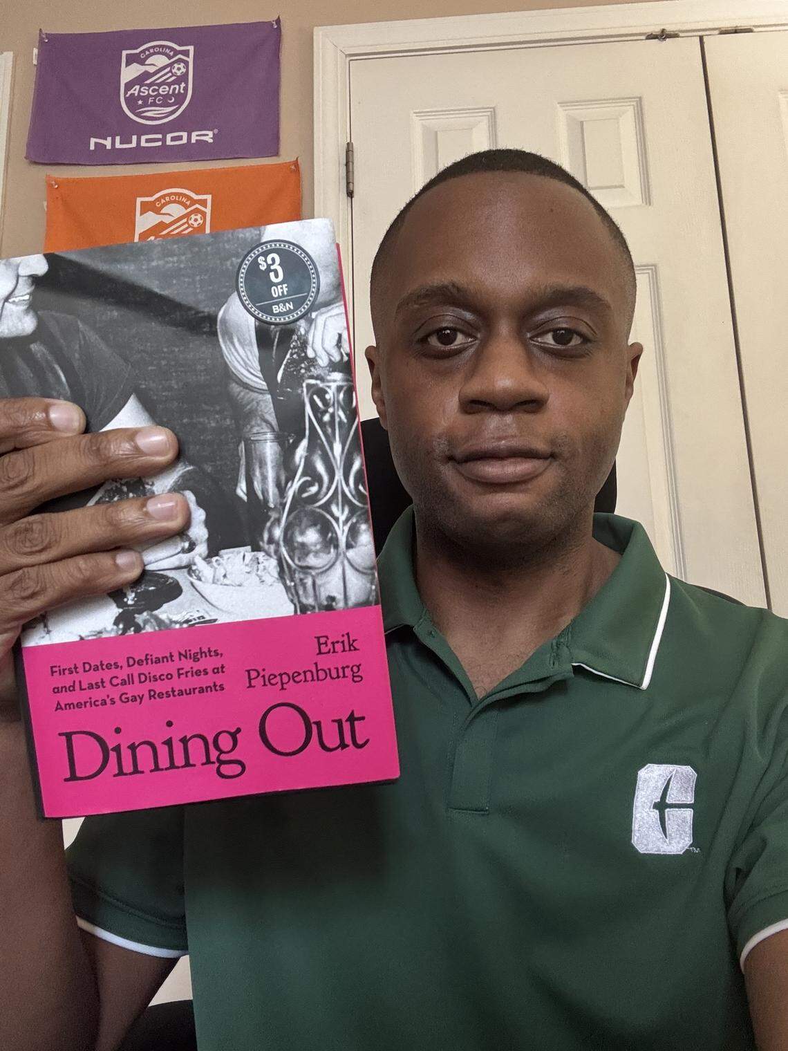 A person in a green polo shirt is holding up a copy of the book “Dining Out” by Erik Piepenburg, posing for a photo. Behind them, three purple and orange flags with the Carolina and Nucor logos hang on the wall.
