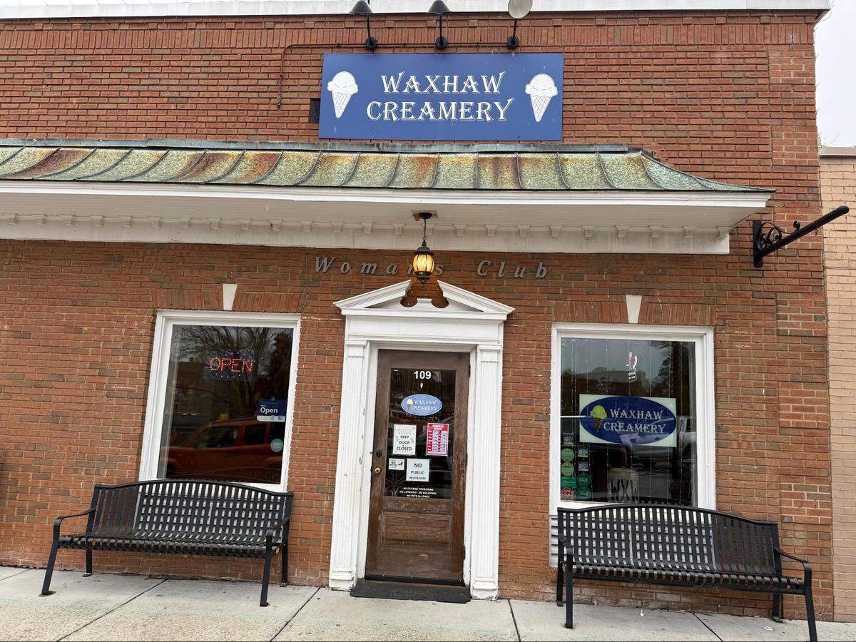 The exterior of a classic red brick building with a blue sign that reads “Waxhaw Creamery” flanked by ice cream cone illustrations. Below the roofline, the words “Woman’s Club” are etched into the brick. Two black benches sit on the sidewalk on either side of a dark wood entrance door.