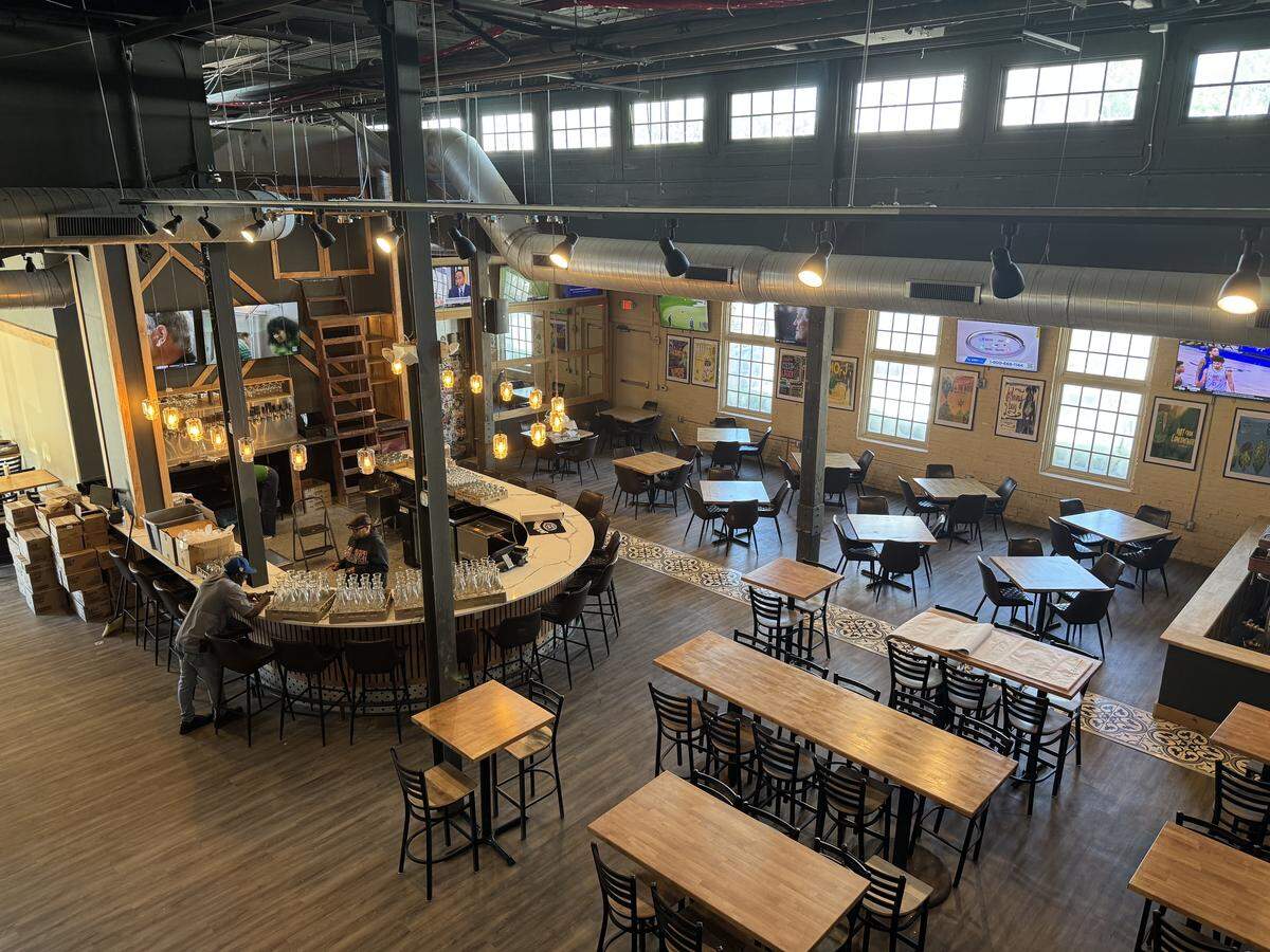 A high-angle, wide interior shot of a spacious, industrial-style brewery and restaurant. The room features high ceilings with exposed ductwork, large windows, and a mix of wooden dining tables and long communal tables. A curved white bar with a wood-slat base is the centerpiece, illuminated by hanging Edison bulb pendant lights. Several flat-screen TVs are mounted on the walls.